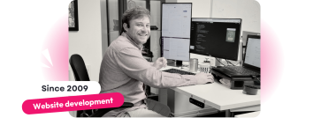 A black-and-white photo of a website development specialist smiling while working at a desk with multiple monitors displaying code and documents